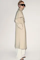Trench de cuello con solapa y manga larga, con detalle de capa desmontable del mismo tejido en la parte superior. Cuenta con cierre frontal de botones y bolsillos en el delantero.