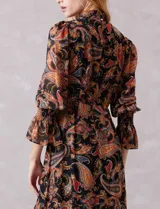 Vestido midi de gasa con estampado de arabescos en tonos cálidos sobre fondo negro. Presenta cuello alto con detalle de punto smock, mangas largas con puños elásticos y volados, y falda con tajo lateral y ruedo con volado. Incluye forro interno y cierre posterior.