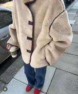 Chaqueta tipo teddy de corte holgado, color beige, con cuello alto, cierre frontal con botones y detalles de ribetes en color marrón oscuro.