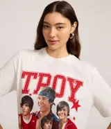 Remera de manga corta color blanco roto con cuello redondo. Presenta un estampado grande en el frente con la palabra "TROY" en letras rojas con textura de puntos blancos, y un collage de cuatro imágenes del actor Zac Efron en su papel de Troy Bolton de High School Musical, rodeado de estrellas rojas.