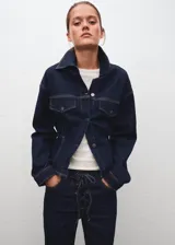 Chaqueta estilo tejano vaquero de algodón con elasticidad, color azul marino. Cuenta con cuello camisero, cierre frontal con botones, dos bolsillos de parche con solapa y botón en el pecho, mangas largas con puños abotonados y tira ajustable en la parte posterior.