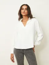 Blusa de color blanco con cuello en V y mangas largas con puños abotonados. Presenta un diseño de corte recto y holgado.