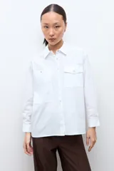 Camisa blanca de manga larga con cuello clásico y botones frontales. Presenta dos bolsillos asimétricos en el pecho, uno con solapa y otro sin.