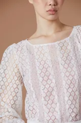 Blusa de encaje blanco con mangas largas abullonadas y puños con detalle de terminación desflecada. Presenta un diseño de cuello redondo y corte recto.