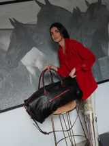Bolso weekender de cuero negro, con cierres metálicos rojos, asas de mano y correa ajustable para hombro en tono marrón.