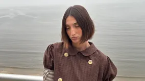 Bolero de corte corto y diseño cruzado con doble botonadura frontal, confeccionado en tela a cuadros color marrón. Presenta cuello redondo y mangas cortas abullonadas.