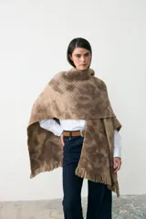 Ruana de tejido de punto con diseño teñido artesanalmente en tonos beige y marrón, con terminación de flecos en el ruedo y etiqueta de cuero sintético en el lateral.