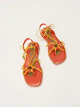 Sandalias planas de cuero color marrón, con diseño de punta cuadrada, tiras finas cruzadas y un detalle trenzado circular en el empeine. Cuentan con tiras para atar al tobillo decoradas con cuentas turquesas en las puntas.