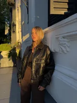 Campera tipo bomber de cuero negro con cuello clásico, cierre frontal metálico y bolsillos frontales con solapa. Presenta un diseño de corte holgado con elástico en la cintura.