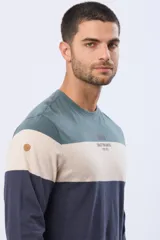 Remera de manga larga con cuello redondo y diseño de rayas horizontales en azul verdoso, beige y azul marino. Presenta una estampa frontal con texto y un parche decorativo en la manga.