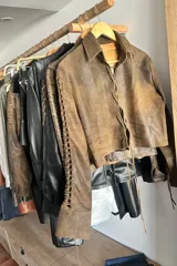 Camisa corta marrón de cuero ovino desgastado, con cuello clásico, cierre frontal con botones, mangas largas con cutouts y puños anchos dorados.