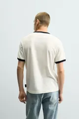 Camiseta de algodón color crudo con cuello y bordes de mangas en contraste negro. Presenta un estampado frontal de estilo vintage con el personaje Striker, mascota de la Copa Mundial de la FIFA 1994, jugando al fútbol.