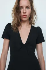 Vestido corto de color negro con escote en pico profundo, mangas cortas abullonadas y detalle de lazo frontal. Presenta un diseño entallado con cierre de cremallera oculto en la espalda.