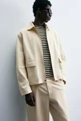 Cazadora de hombre color beige claro, corte relajado, con cuello solapa, manga larga y puños con botones a presión. Presenta bolsillos de plastrón con solapa en la cadera y cierre frontal de cremallera oculta por solapa de botones a presión.