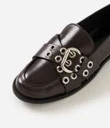 Mocasín Satinato de poliuretano color marrón oscuro, con punta redondeada, tacón bajo y empeine decorado con tiras cruzadas, hebilla ajustable y ojetes metálicos.