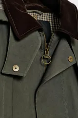 Trench de estilo encerado color verde militar con cuello de solapa en pana marrón, capucha extraíble y capa superior. Presenta cierre frontal con cremallera y botones a presión ocultos, bolsillos delanteros de plastrón con solapa y mangas largas con trabillas.