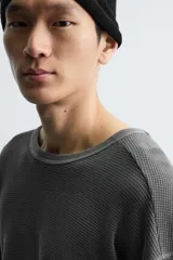 Camiseta de manga larga con cuello redondo, confeccionada en tejido de algodón con textura tipo waffle y efecto lavado. Presenta un corte relajado con puños en rib y bajo irregular.
