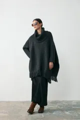 Poncho de tejido artesanal en color negro, con cuello alto tipo polera, mangas largas y terminación con flecos en el ruedo. Presenta una etiqueta de cuero decorativa en la parte inferior.