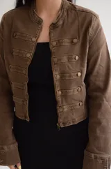 Chaqueta de estilo militar en color marrón, con corte estructurado y cuello mao. Presenta cierre frontal con cremallera y detalles de filas horizontales con botones metálicos decorativos en el frente y en los puños.
