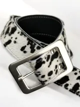 Cinturón de cuero genuino con estampado de manchas tipo vaca en blanco y negro, con hebilla rectangular de metal plateado.
