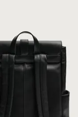 Mochila de diseño minimalista en color negro, confeccionada en material sintético con acabado liso. Presenta una solapa frontal con cierre de hebilla decorativa y sistema de imán, compartimento principal con cremallera, bolsillos laterales externos, asa superior de mano y correas de hombro ajustables y acolchadas.