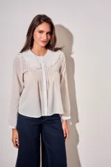 Blusa blanca de algodón peinado con mangas largas, cuello babero bordado y pliegues frontales.