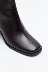 Bota corta de caña elástica, color negro con acabado charolado. Presenta puntera cuadrada y taco medio geométrico y ancho.