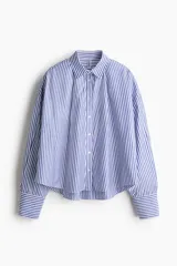 Camisa de corte holgado con estampado de rayas verticales azules y blancas. Presenta cuello clásico, cierre frontal con botones, hombros caídos y mangas largas con puños abotonados. El diseño incluye un canesú con pliegue en la espalda y bajo redondeado con la parte posterior ligeramente más larga.