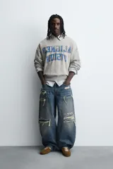 Sudadera gris de corte boxy fit, con cuello redondo y manga larga. Presenta un estampado frontal con texto en azul con efecto desgastado y acabados en rib en puños y ruedo.