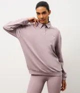 Buzo deportivo de corte oversized color lila, con cuello polo y cierre frontal con botones a presión. Presenta mangas largas con puños ajustados y una estampa bordada en el pecho. Confeccionado en una mezcla de poliéster, modal y elastano.