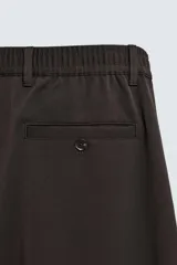 Pantalón de corte recto confeccionado en tejido elástico, con cintura elástica, pliegues frontales, bolsillos laterales y bolsillos traseros de vivo con botón.