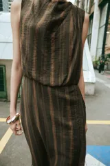 Vestido midi sin mangas de cuello redondo, con diseño de rayas verticales en tonos marrones. Presenta un detalle drapeado en el frente, volantes laterales y un bajo asimétrico. Cierre posterior con cremallera oculta.