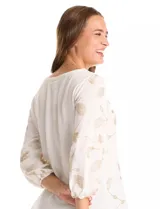 Blusa de tejido fino color marrón con bordado floral en hilo dorado. Presenta cuello redondo, mangas tres cuartos abullonadas con puños elásticos fruncidos y ruedo redondeado con corte asimétrico.