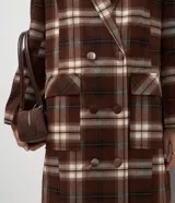 Trench coat largo de corte clásico con estampado de cuadros en tonos marrones y beige. Presenta cuello con solapas, cierre frontal con botones, mangas largas y bolsillos laterales aplicados.