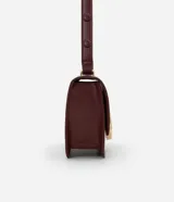 Cartera tipo shoulder bag de tamaño mediano, confeccionada en poliuretano color marrón oscuro. Presenta una solapa frontal con un detalle metálico dorado de forma orgánica y asa ajustable.