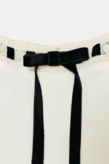 Camisa de georgette color blanco con escote cuadrado, mangas largas acampanadas y forro interior. Presenta detalles de puntilla a tono en el escote y un lazo de terciopelo negro en el centro del pecho.