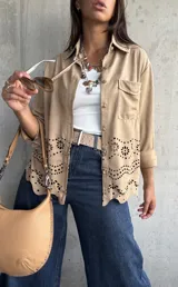 Sobrecamisa de gamuza color beige, con cuello camisero, cierre frontal con botones metálicos y bolsillo en el pecho. Presenta un diseño con calados florales y bordes ondulados en la parte inferior.