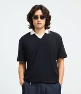 Remera de corte boxy en tejido crepé, color negro, con cuello polo en contraste color blanco y mangas cortas. Presenta un diseño holgado y moderno con textura sutil en la superficie.
