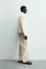 Cazadora de hombre color beige claro, corte relajado, con cuello solapa, manga larga y puños con botones a presión. Presenta bolsillos de plastrón con solapa en la cadera y cierre frontal de cremallera oculta por solapa de botones a presión.