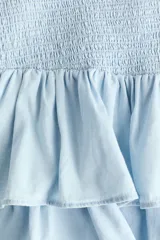 Pollera mini confeccionada en denim de algodón celeste, con cuerpo ajustado en punto smock y doble volante al bies en el bajo.