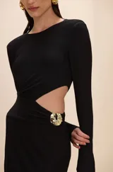 Vestido corto negro de manga larga, con diseño cut-out lateral en la cintura y detalle de aplique metálico dorado en la cadera. Presenta cuello redondo y silueta ajustada.