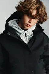 Anorak corto de color negro con acolchado interior, cuello subido y capucha. Presenta mangas largas con puños elásticos, bolsillos delanteros con cierre y bajo ajustable con cordones. Cierre frontal con cremallera termosellada y botones a presión.