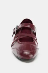 Bailarinas color bordó con efecto acharolado, detalle de pespuntes, cierre de hebilla metálica ajustable y tiras elásticas cruzadas en la parte frontal. Acabado en punta redonda y suela de 0,5 cm.