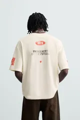 Camiseta de manga corta color crema, corte boxy fit, con cuello redondo. Presenta un estampado frontal en negro con el texto 'VORTEX PULSE' y el número '1974' en un óvalo rojo desgastado. Tiene pequeños detalles gráficos rojos en las mangas.