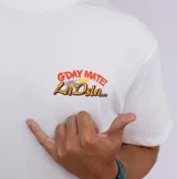Remera blanca de manga corta con estampado frontal que dice "G'DAY MATE! La Isla..." en colores naranja y amarillo.