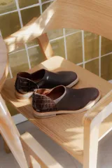 Zapatos tipo mocasín de cuero color marrón oscuro, con diseño de textura lisa en la parte delantera y textura de reptil en el talón. Cuentan con cierre trasero y suela plana de color claro.