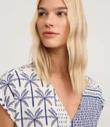 Enterito corto de viscosa y lino color blanco con estampado tropical en azul de palmeras y soles, escote en V, manga corta caída, bolsillos laterales y cinto de cuerda trenzada en la cintura.