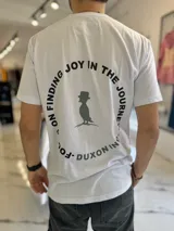 Remera blanca de manga corta con estampa en la espalda que incluye la frase 'Focus on finding joy in the journey' formando un círculo alrededor de la figura de un pájaro con sombrero.