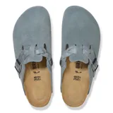 Zueco tipo mule de gamuza color gris, con diseño de tira trenzada sobre el empeine y hebilla metálica ajustable. Cuenta con plantilla de corcho anatómica y suela de goma negra.