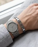 Pulsera masculina con diseño de eslabón central de acero inoxidable con patrón geométrico griego, flanqueado por secciones de acero y correa de cuero gris.